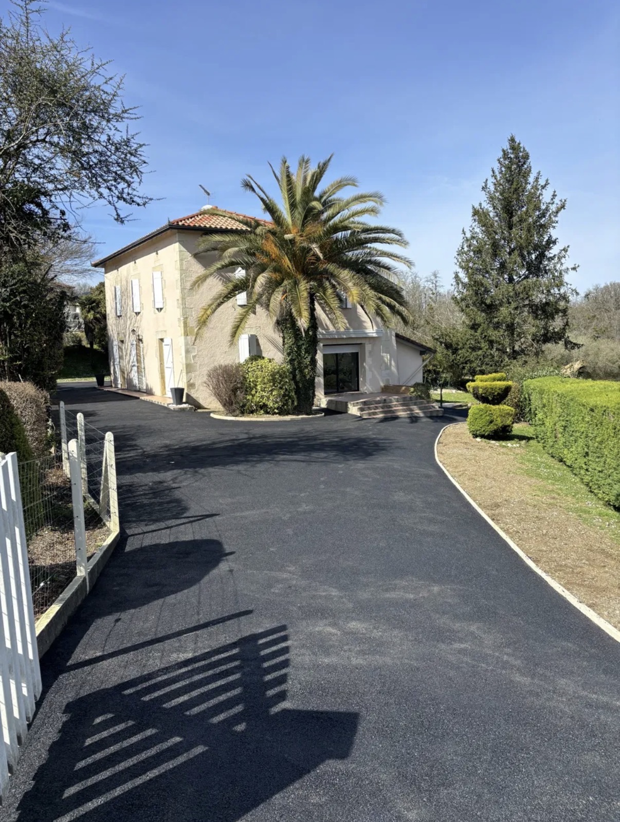 Grande cour en enrobé noir avec bordures blanches courbes devant une maison landaise