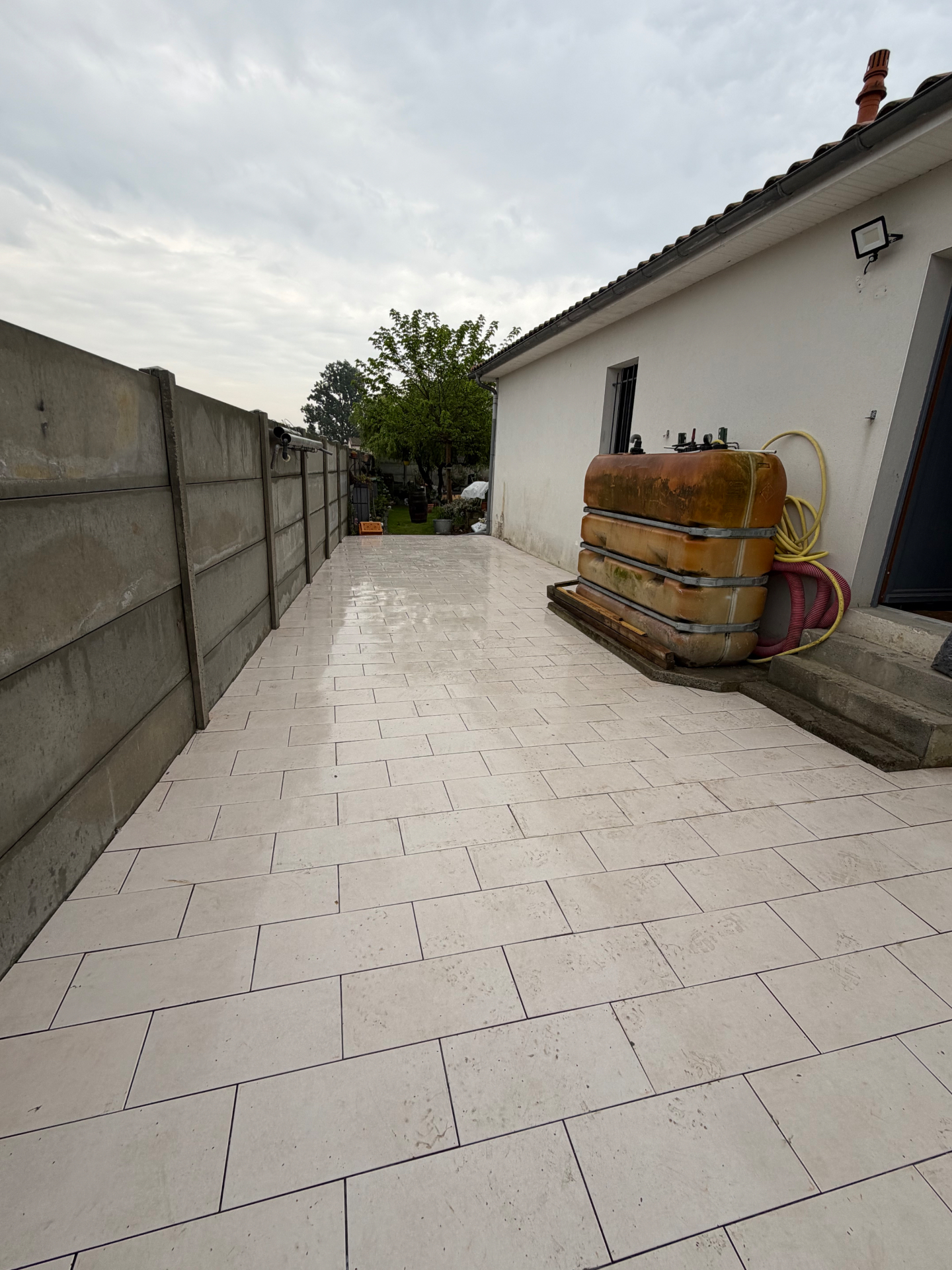 Terrasse latérale terminée en dallage clair posé entre la façade de la maison et le mur de clôture en béton