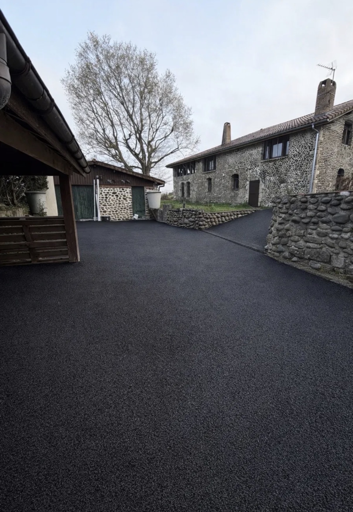 Cour et allée de garage en enrobé noir devant une bâtisse en galets, avec abri voiture en bois et murets de galets
